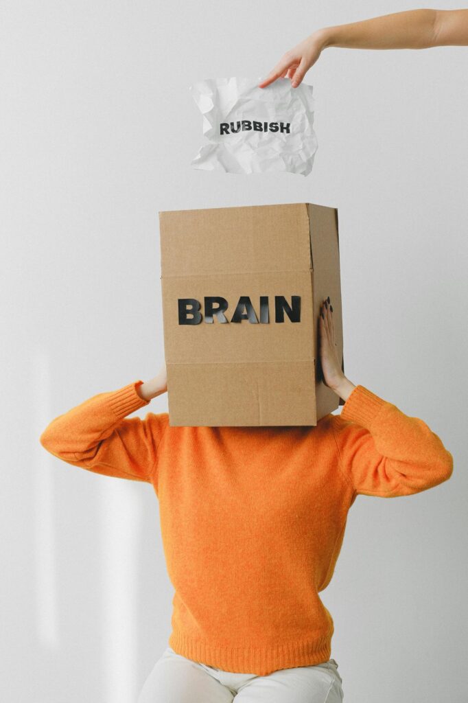 A man puts his head in a box. Brain is written on the top of the box. And from above, a man is writing a paper abbreviated and throwing it into the box.